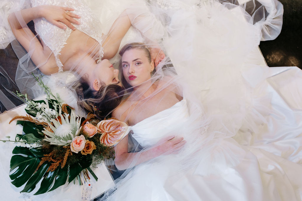 Two brides lying on a black and white chequered dance floor with a veil draped over them, creating a dreamy, romantic bridal editorial scene