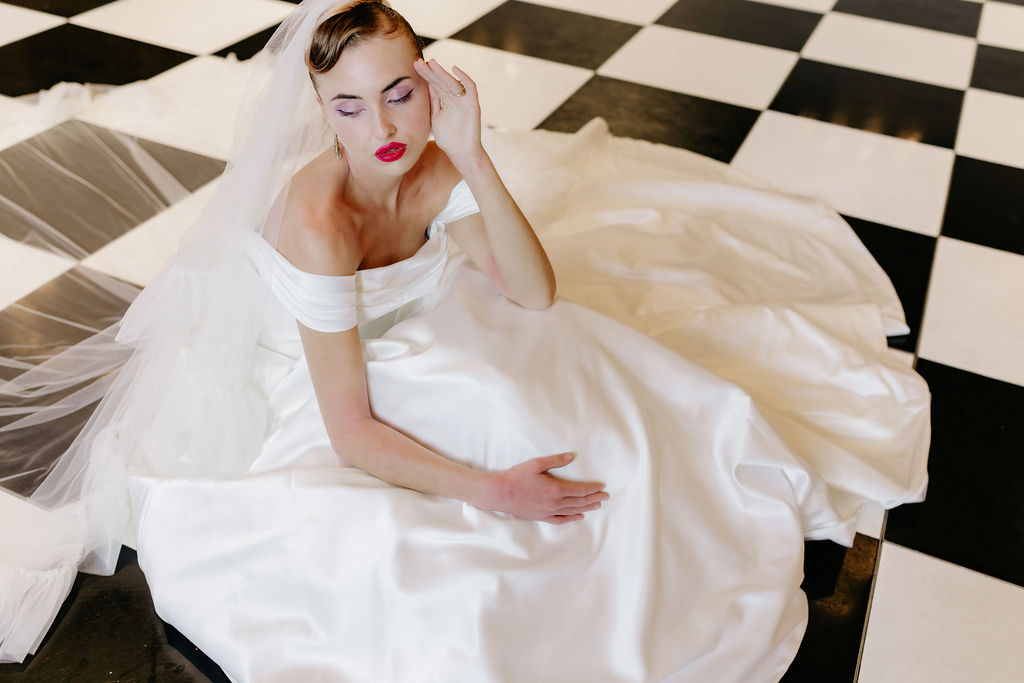 Bridal model sitting on a black and white chequered dancefloor in a modern Madi Lane gown, styled with bold makeup and soft waves